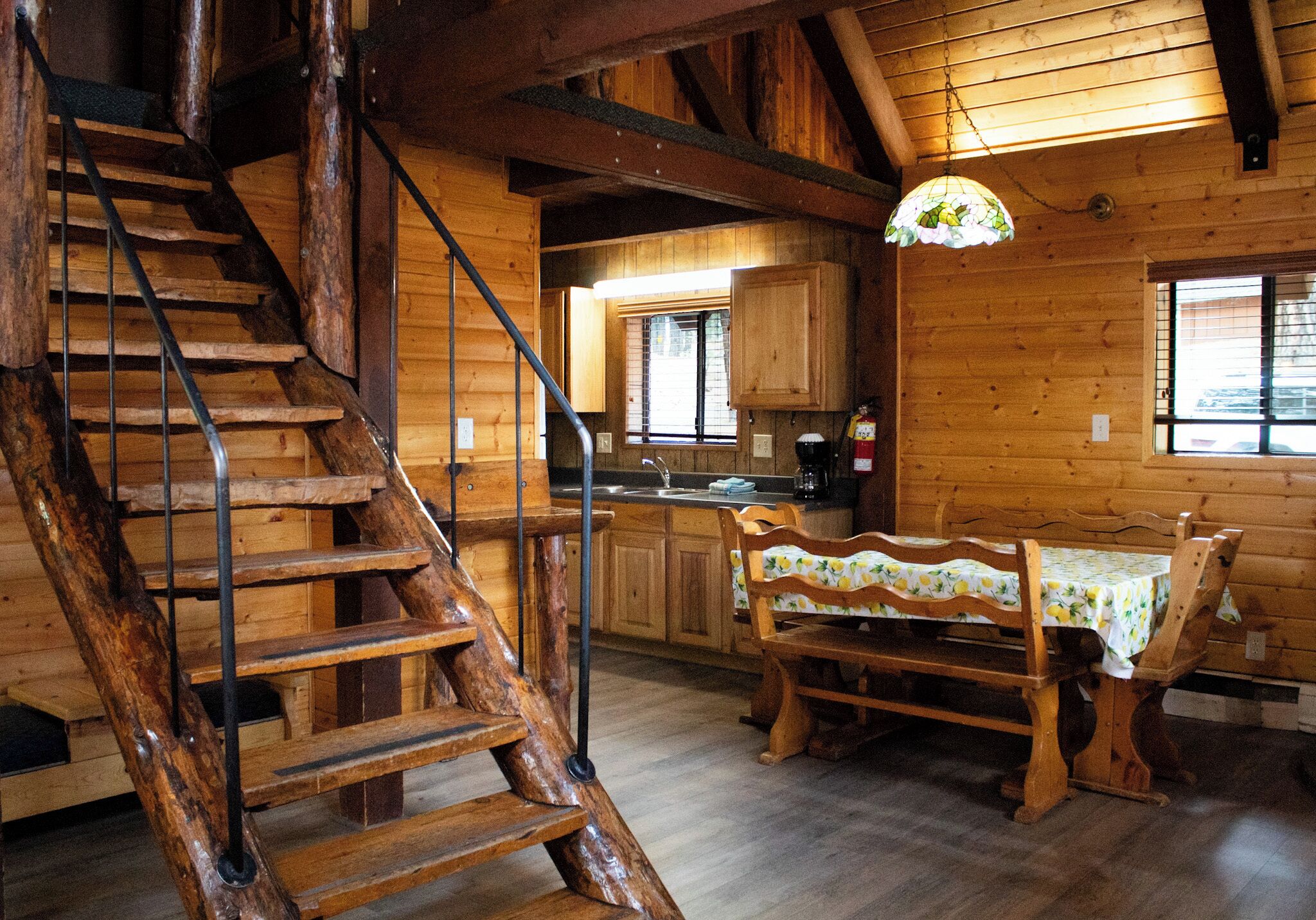 Interior de una cabaña de madera con escalera de madera, una mesa de comedor con sillas y una zona de cocina.
