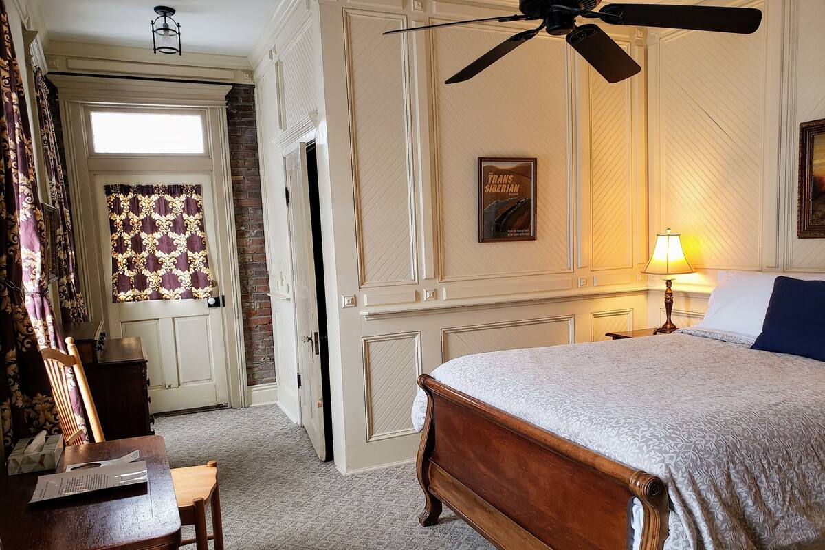Ett hotellrum med en säng i trä, ett skrivbord, en stol, en takfläkt, en lampa och en inramad tavla på väggen.