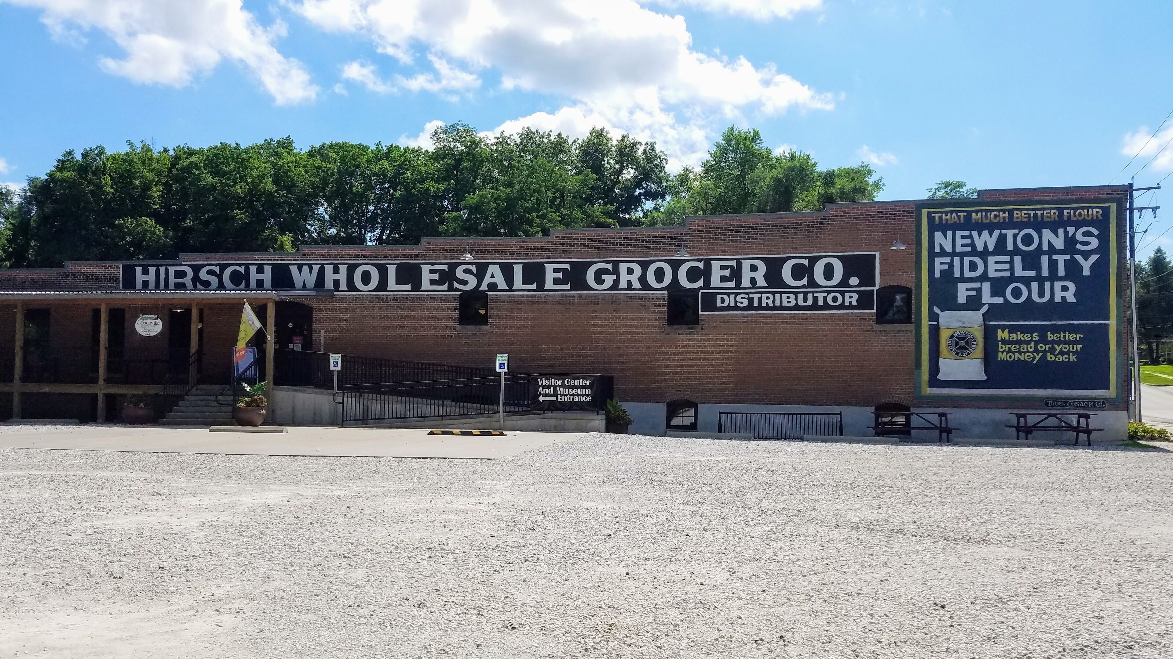 Un bâtiment en briques portant l’inscription « HIRSCH WHOLESALE GROCER CO. DISTRIBUTOR », avec une enseigne indiquant « NEWTON’S FIDELITY FLOUR » et l’entrée d’un centre d’accueil pour les visiteurs.