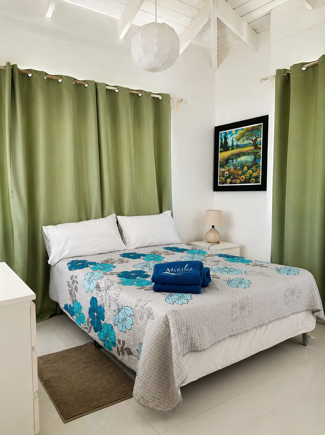 Een slaapkamer met een bed, groene gordijnen, een schilderij, een lamp en een opgerolde handdoek op het bed.