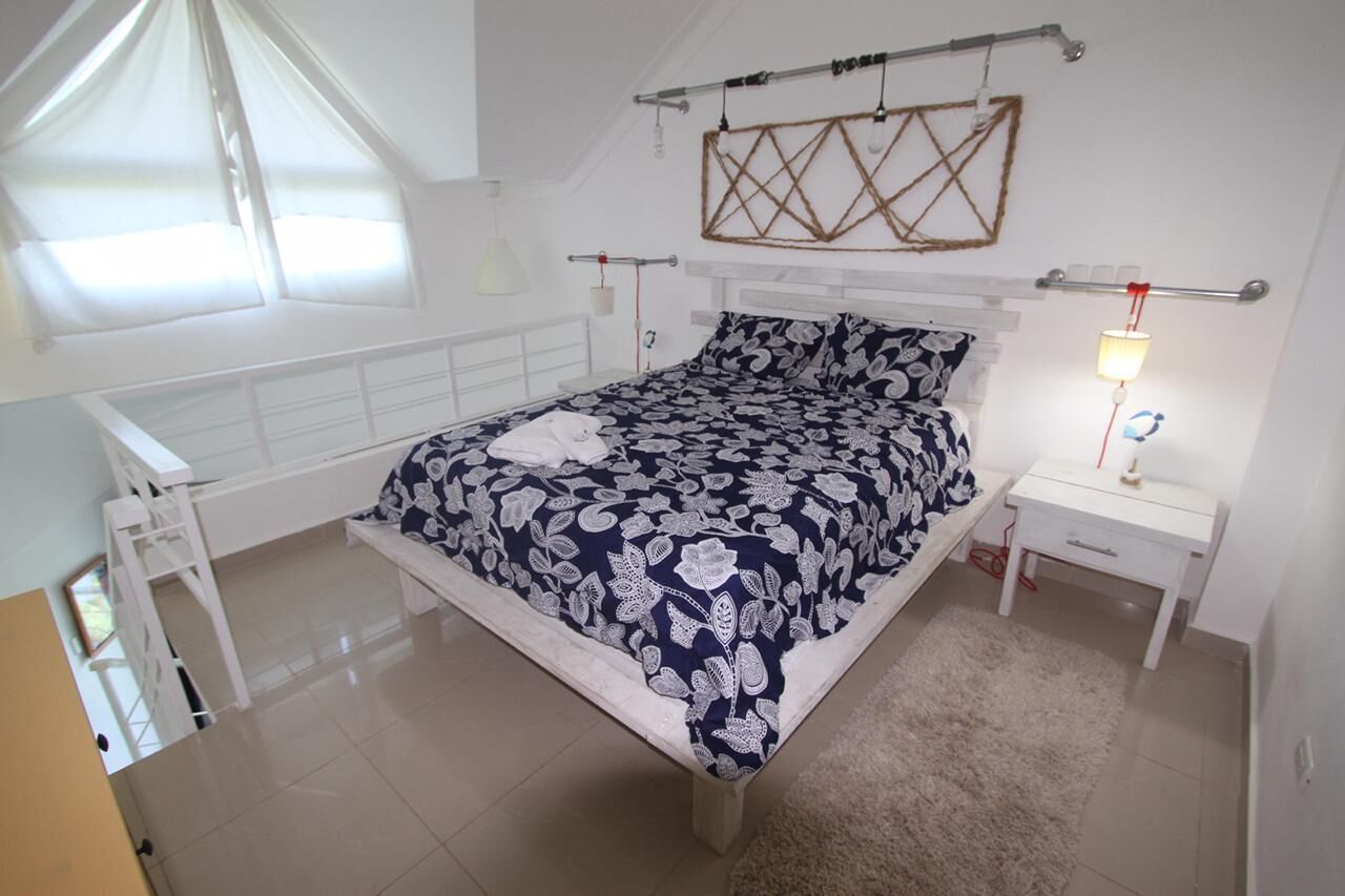 A bedroom with a large bed, a bedside table, a nightstand, a window with curtains, and a wall-mounted light fixture.