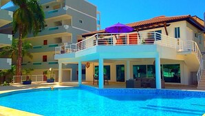 Une piscine dotée d’une zone couverte, de palmiers et d’un immeuble à plusieurs étages avec des balcons.