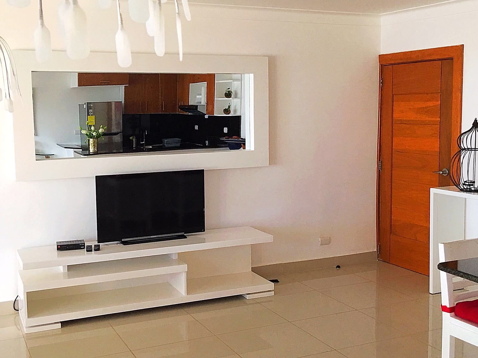 A modern living room with a flat-screen TV on a white entertainment unit, a large mirror above it, and a wooden door to the right.