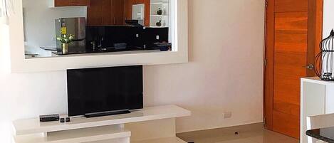 A modern living room with a flat-screen TV on a white entertainment unit, a large mirror above it, and a wooden door to the right.