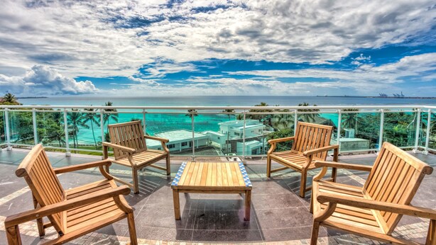 Un balcon avec des chaises et une table en bois, d’où l’on peut admirer une mer turquoise et un paysage urbain lointain.
