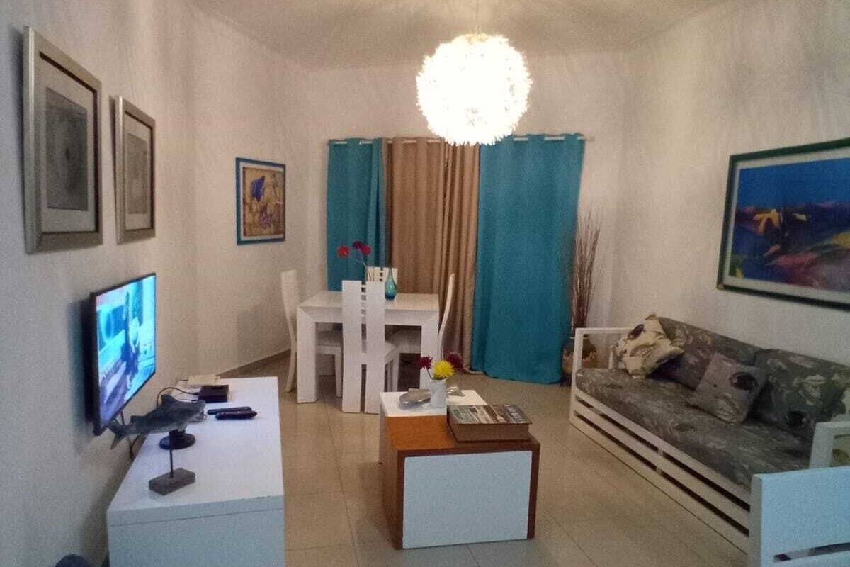Una sala de estar con televisor, mesa de centro, sofá, mesa de comedor con sillas y una araña.