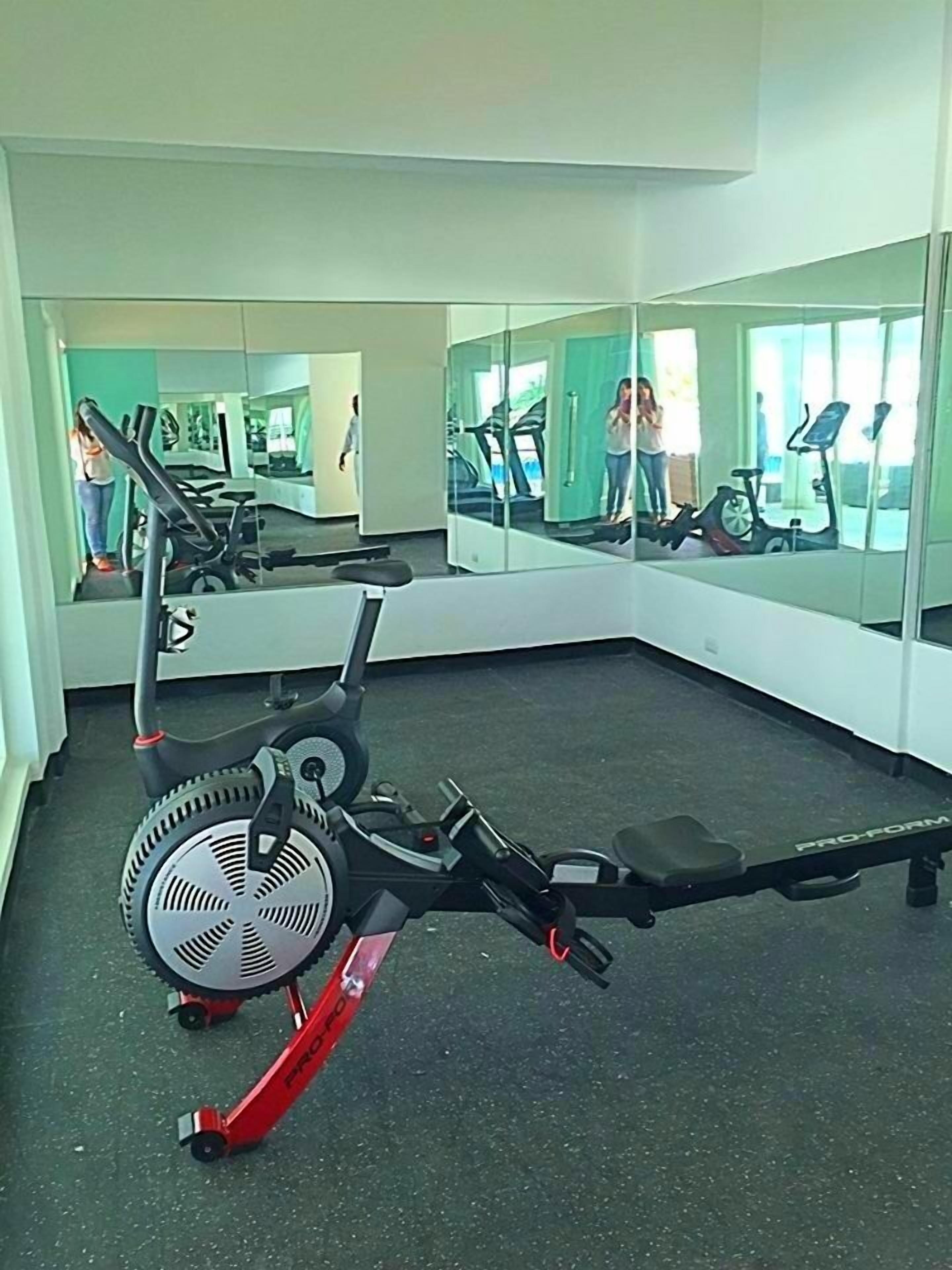 Un gimnasio con equipo de cardio, incluyendo una máquina elíptica y una bicicleta estática, y un espejo grande que refleja el espacio.