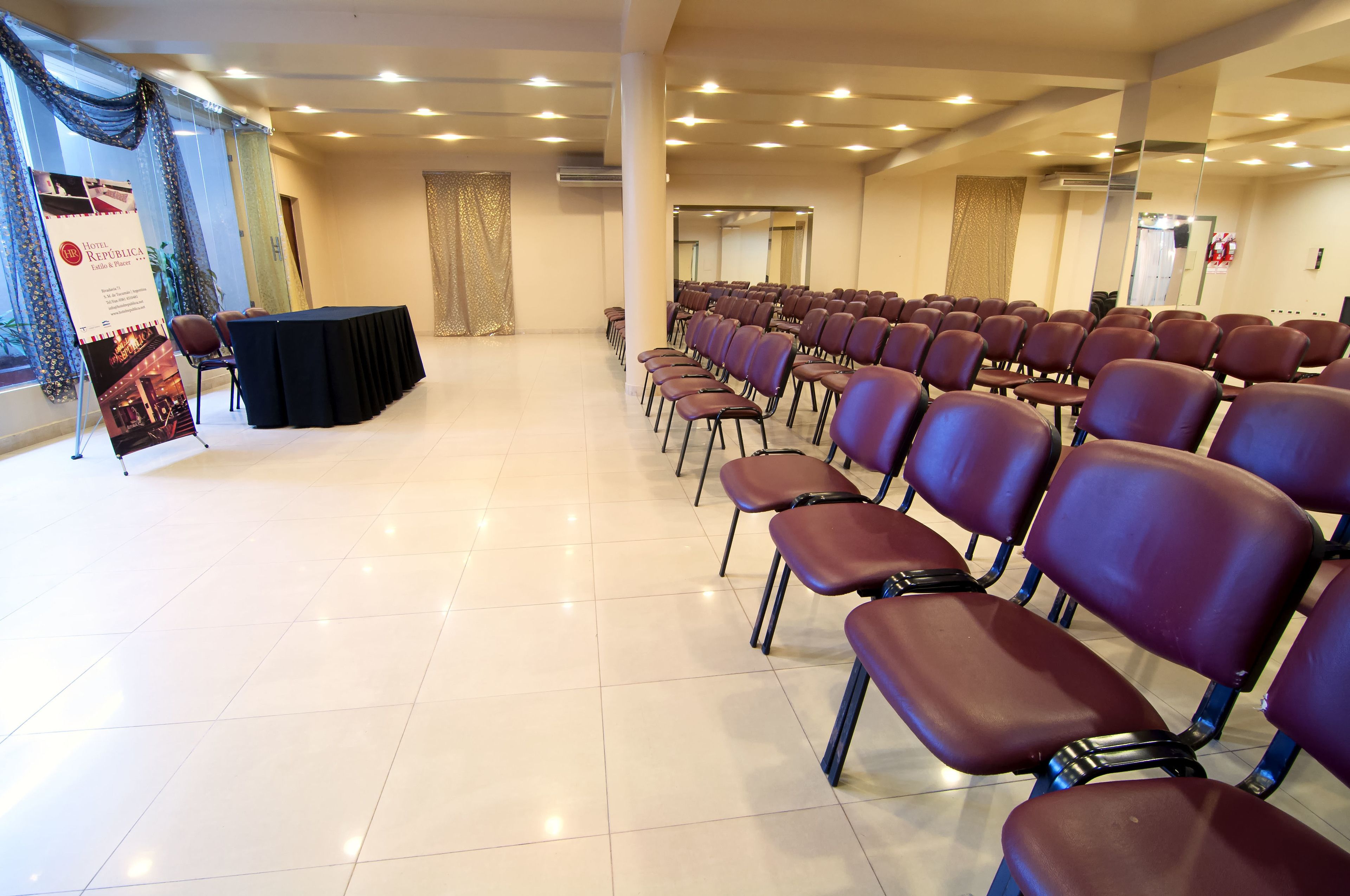 Uma sala de conferências com fileiras de cadeiras, uma mesa preta e um banner à esquerda.