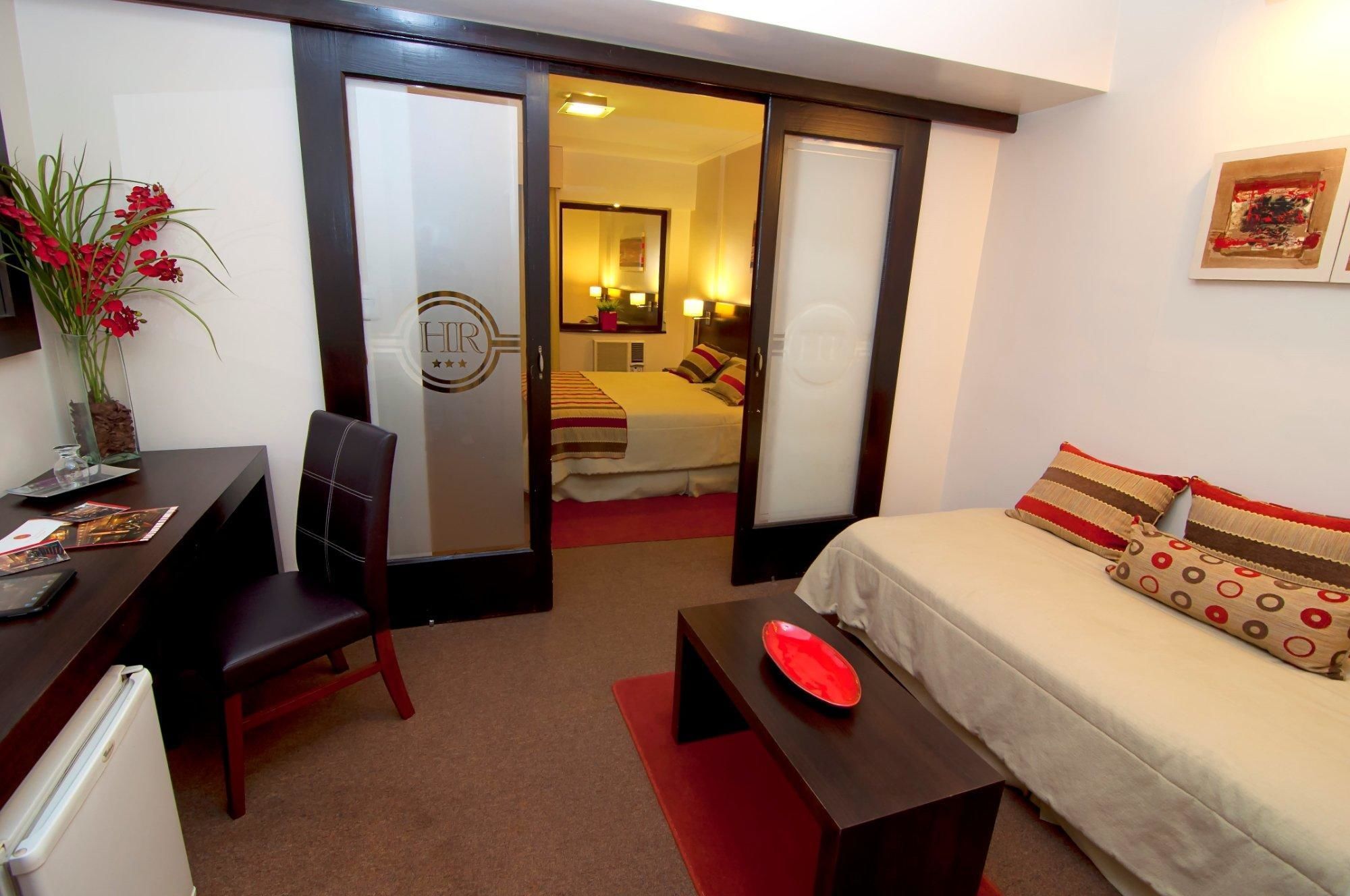 Een hotelkamer met een bed, een bureau, een stoel, een kleine tafel met een rode kom en een potplant.