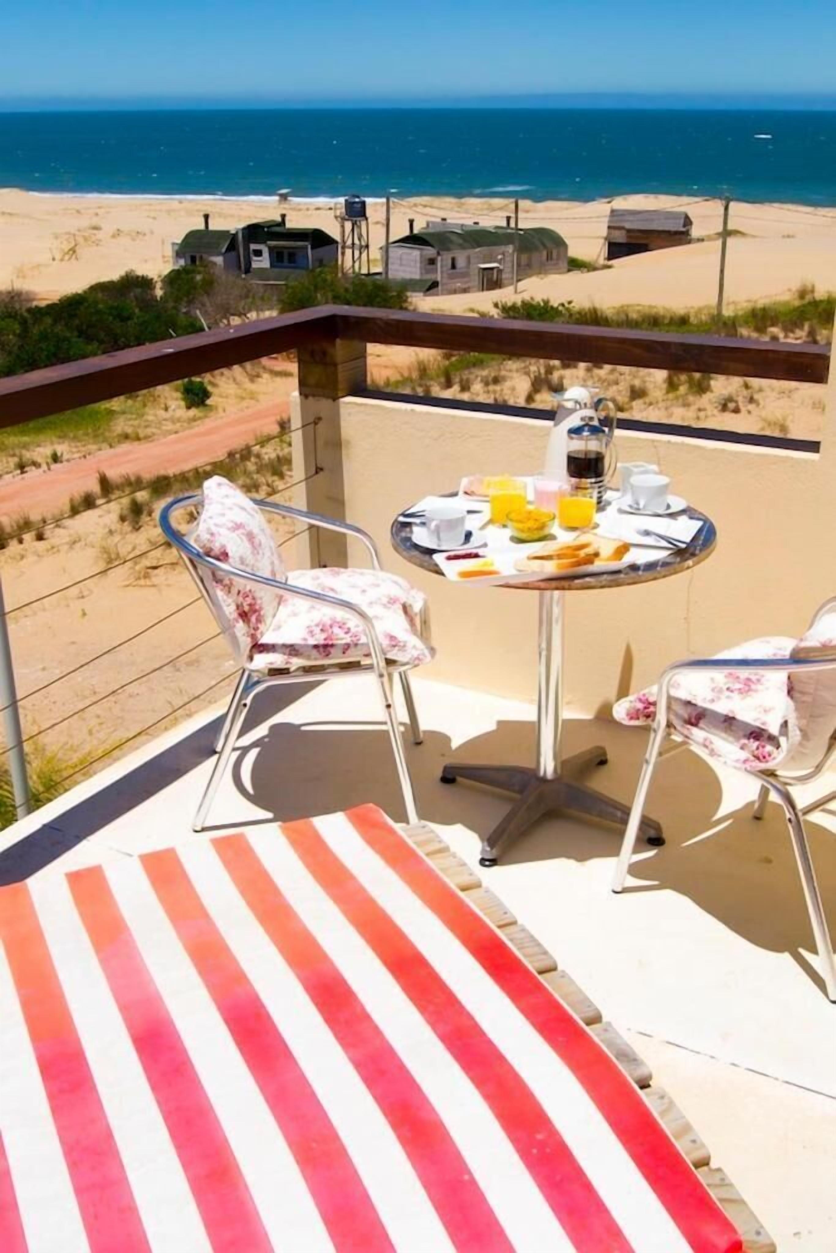 Un balcón con dos sillas y una mesa dispuesta para el desayuno, con vista a una playa y al océano.