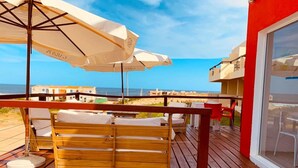En träveranda med utomhusmöbler, vita parasoller och utsikt över stranden och havet.