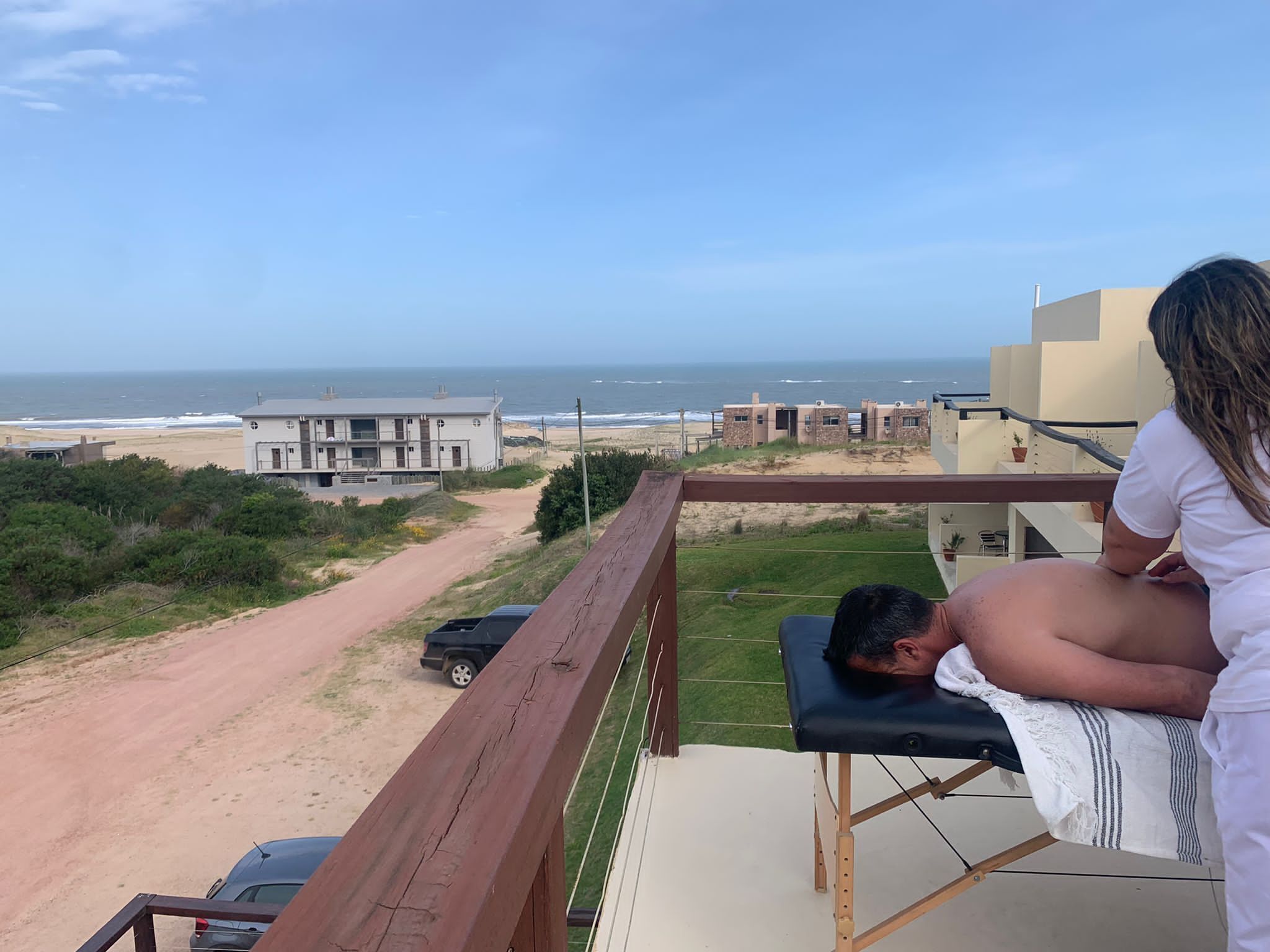 A balcony with a view of the beach and ocean, a massage table, and a person receiving a massage.