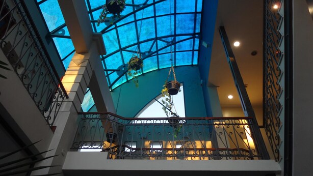 Een meerlaags atrium met een glazen plafond, metalen leuningen en hangende plantenbakken.