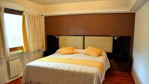 A bedroom with a double bed, two bedside tables, a window with curtains, and a radiator. - Cumbres Hotel (El Bolson)