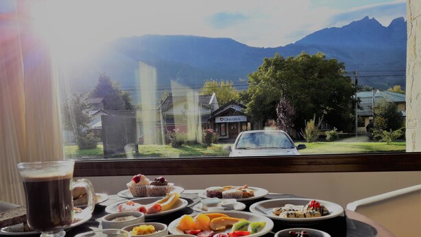 A breakfast table with various dishes and a glass of orange juice, set against a view of mountains and a car in the background. - Cumbres Hotel (El Bolson)