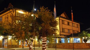 A hotel with a traditional architectural style, featuring a stone facade and multiple windows. There are palm trees wrapped with lights, and flags are visible in front of the building. - Cumbres Hotel (El Bolson)
