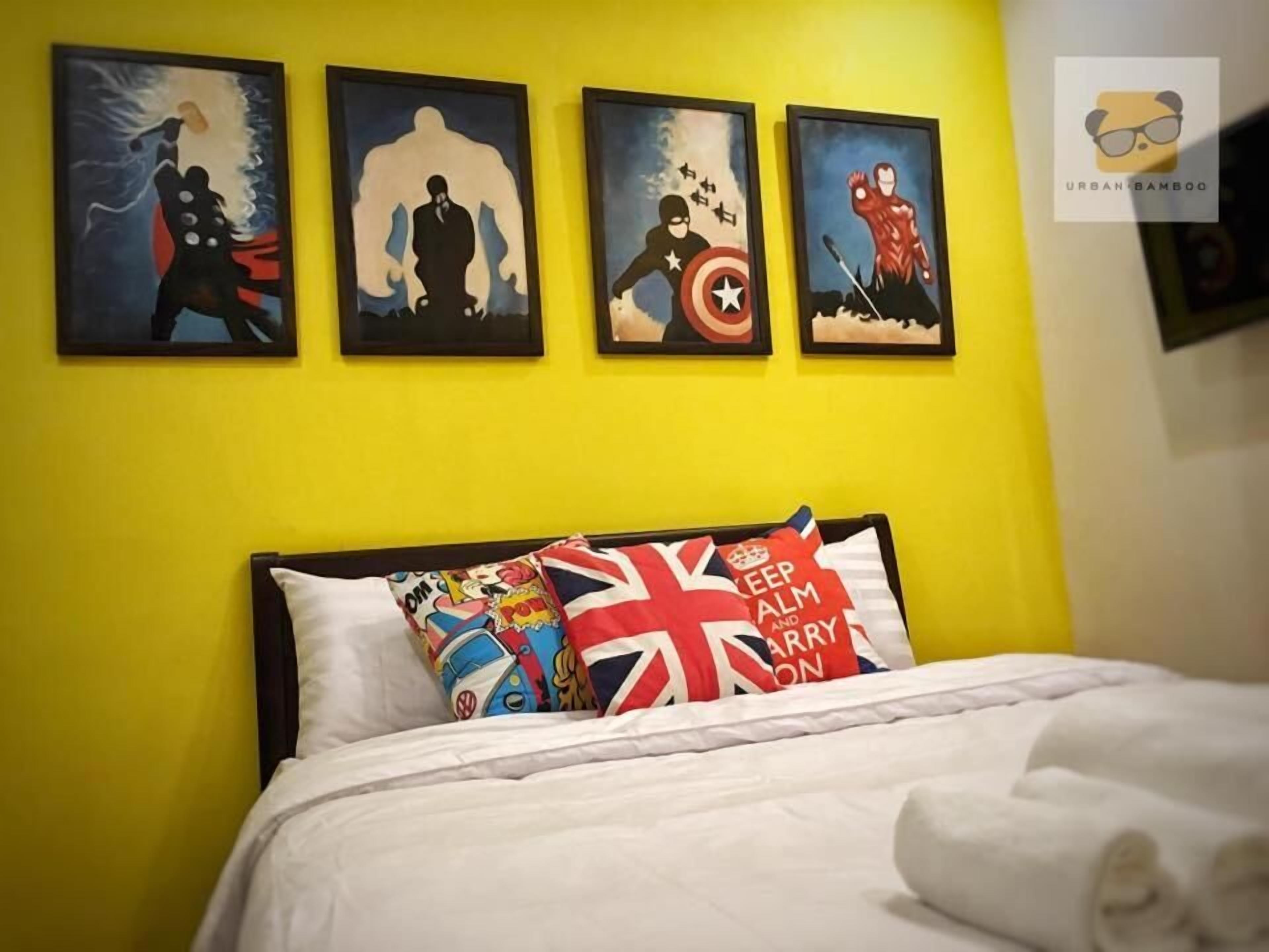 A room with a yellow wall and four framed superhero paintings. There is a bed with white bedding and pillows featuring the Union Jack and a 'Keep Calm and Carry On' design.