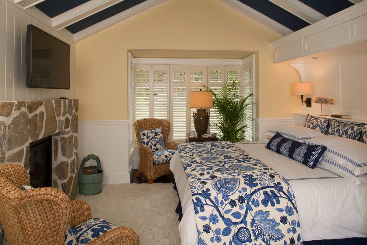 A bedroom with a stone fireplace, a wicker chair, a bed with blue floral bedding, a nightstand with a lamp, and a window with blinds.