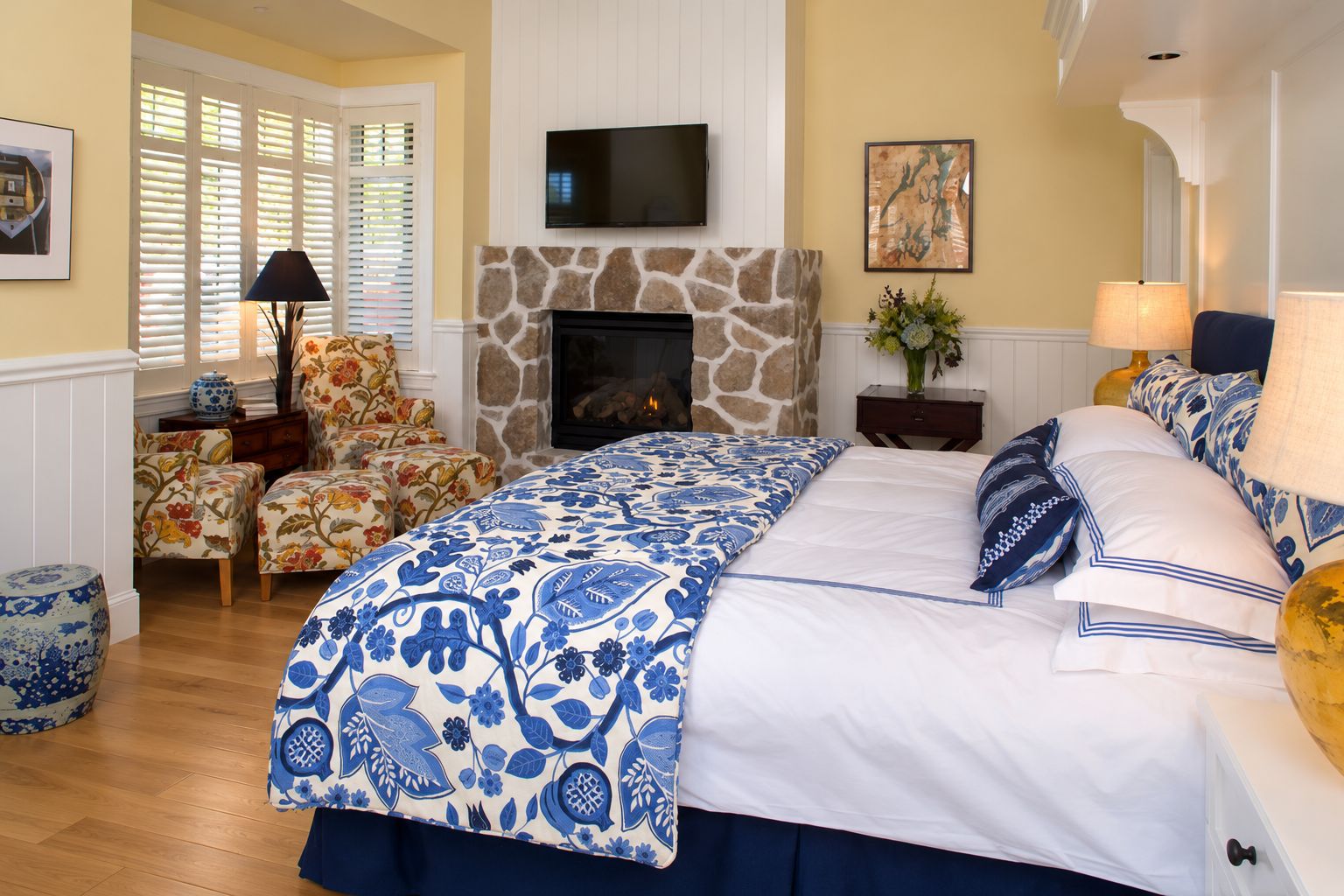 Une chambre avec un grand lit, une cheminée en pierre, un téléviseur placé au-dessus de la cheminée, un fauteuil à motifs floraux et une fenêtre avec des stores.