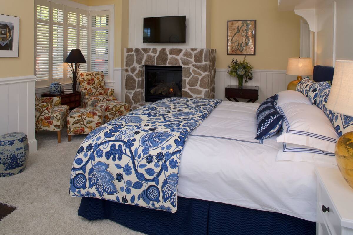 A bedroom with a stone fireplace, a large bed with blue and white bedding, a television mounted above the fireplace, and a window with blinds.