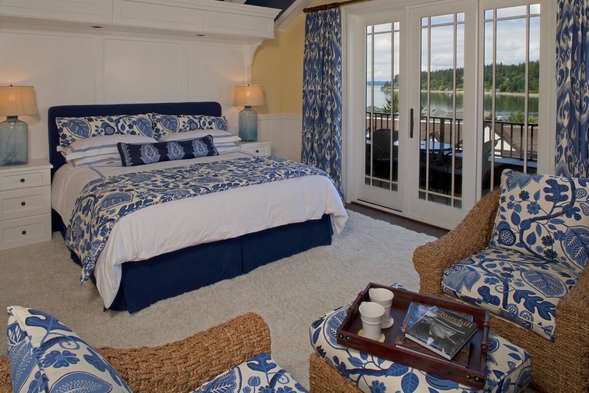 A bedroom with a large bed, a sofa, and a chair, all with blue floral patterns. There is a balcony with a view of the water.