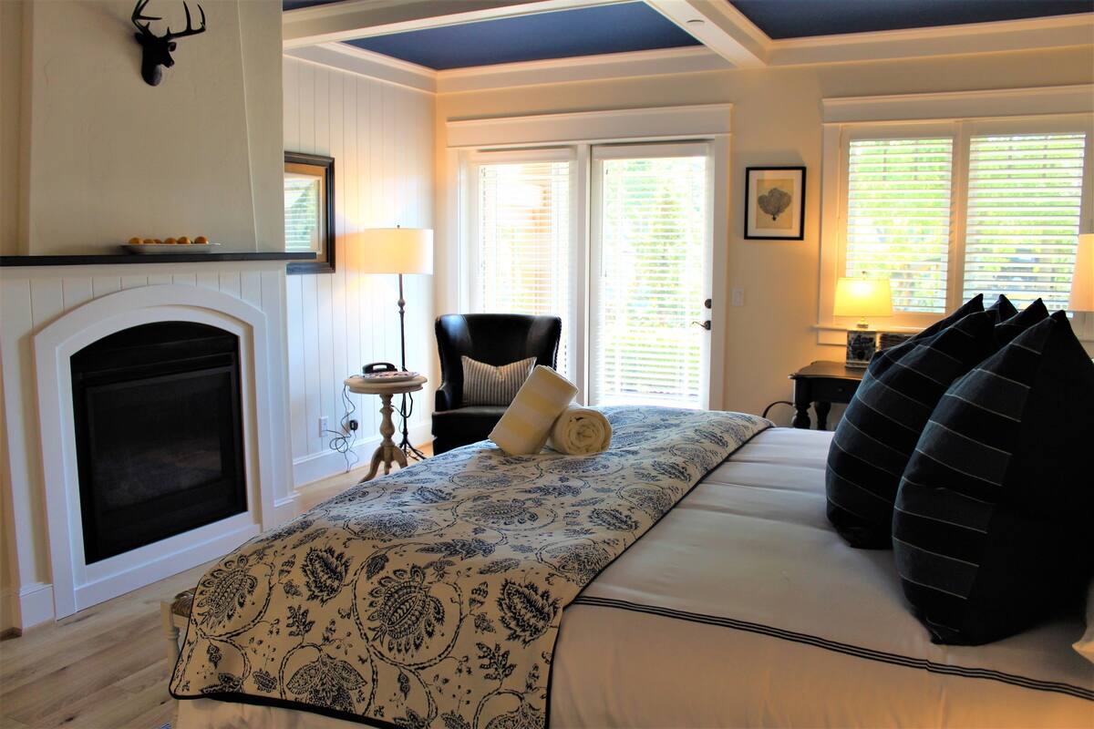 A bedroom with a fireplace, a bed with patterned bedding, a chair, and a lamp.