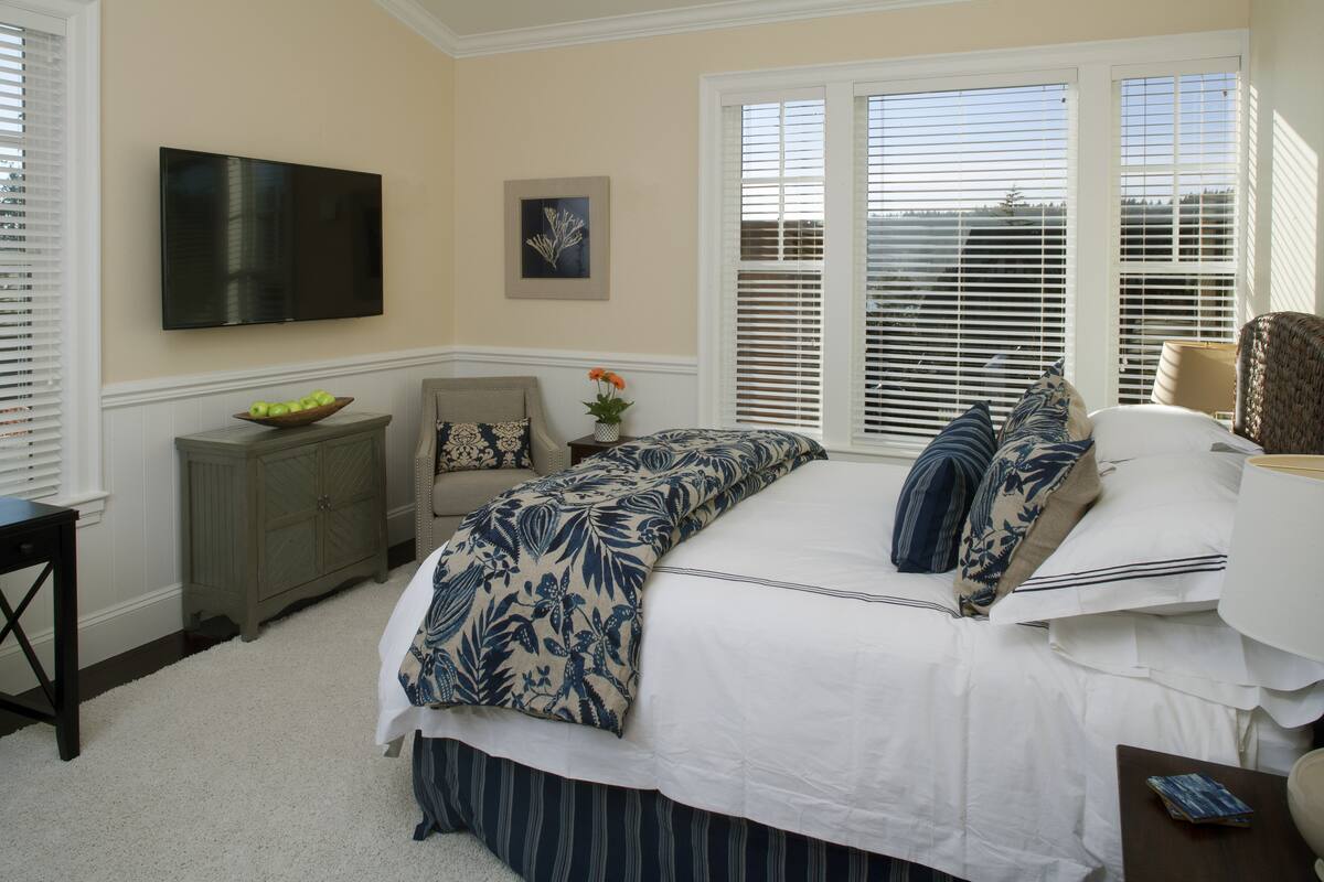 A bedroom with a large bed, a TV mounted on the wall, a window with blinds, a dresser, and a small table with a vase.