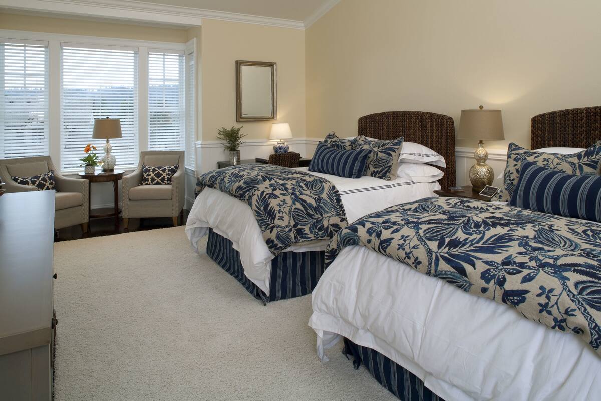 A hotel room with two beds, a sitting area with a chair, a small table with a lamp, and a window with blinds.