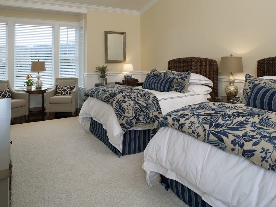 A hotel room with two beds, a sitting area with a chair, a small table with a lamp, and a window with blinds.