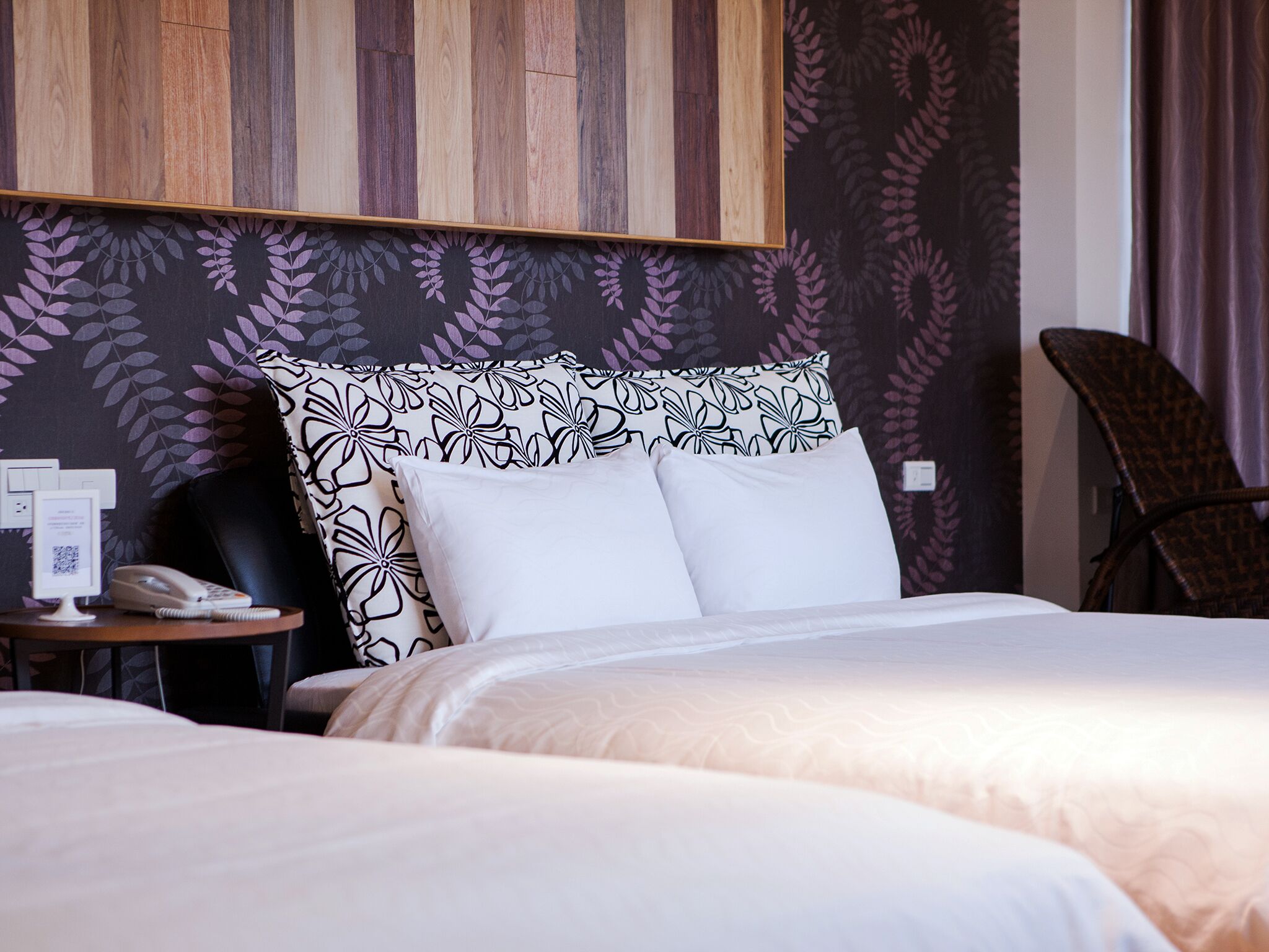 Habitación de hotel con dos camas, cabecero de madera, pared estampada, una mesita con un teléfono y una silla.
