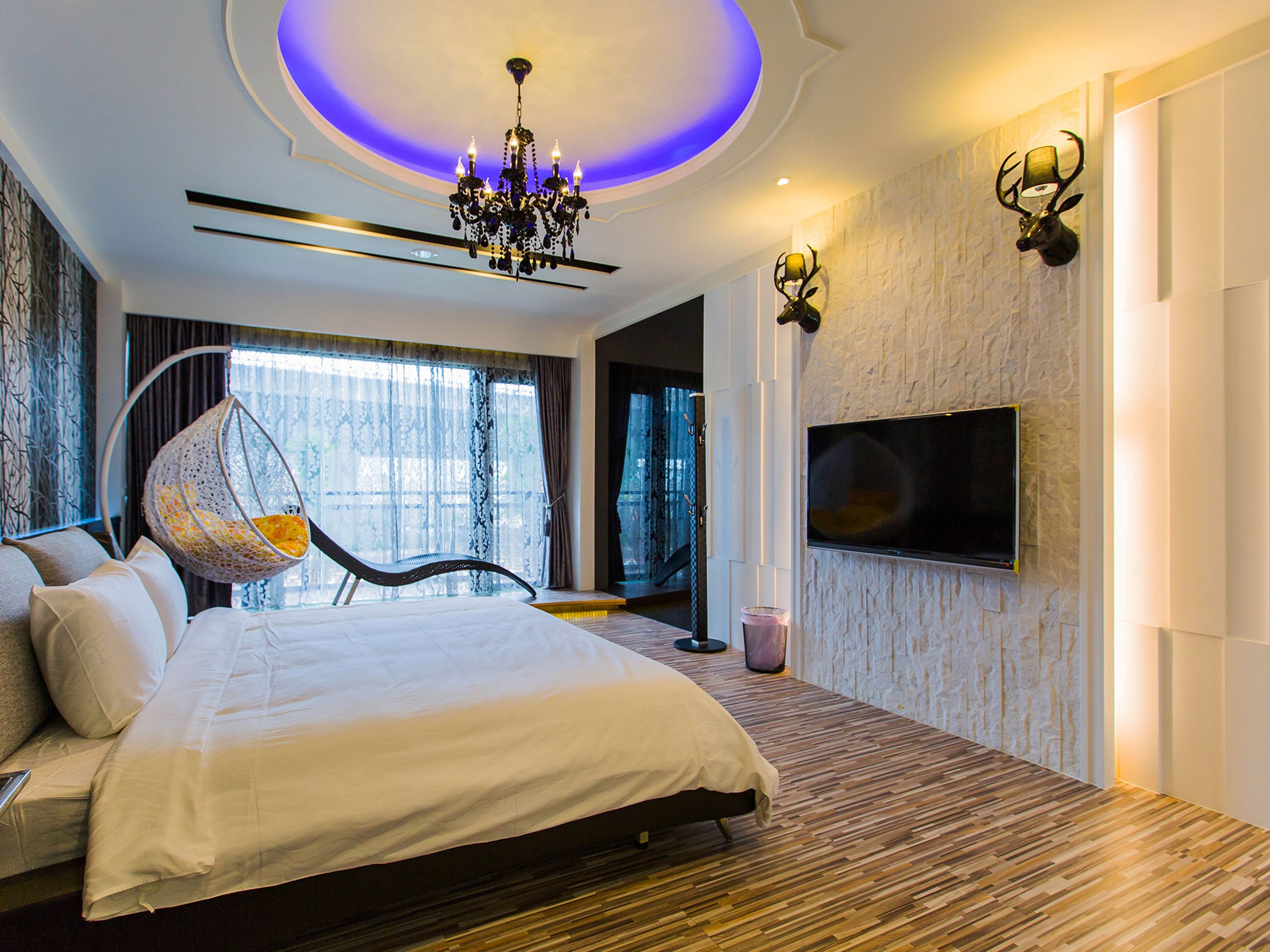 A modern bedroom with a large bed, a hanging chair, a TV mounted on the wall, and a chandelier.