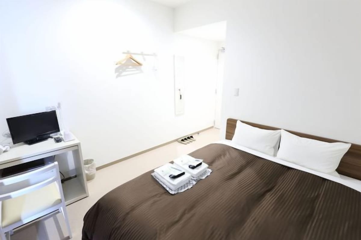 A hotel room with a bed, a desk with a television, a chair, and a small table.
