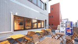 An outdoor seating area with wooden tables and chairs, a vending machine, and a building with large windows and tiled walls. - Oak Hotel Edo (Tokyo)