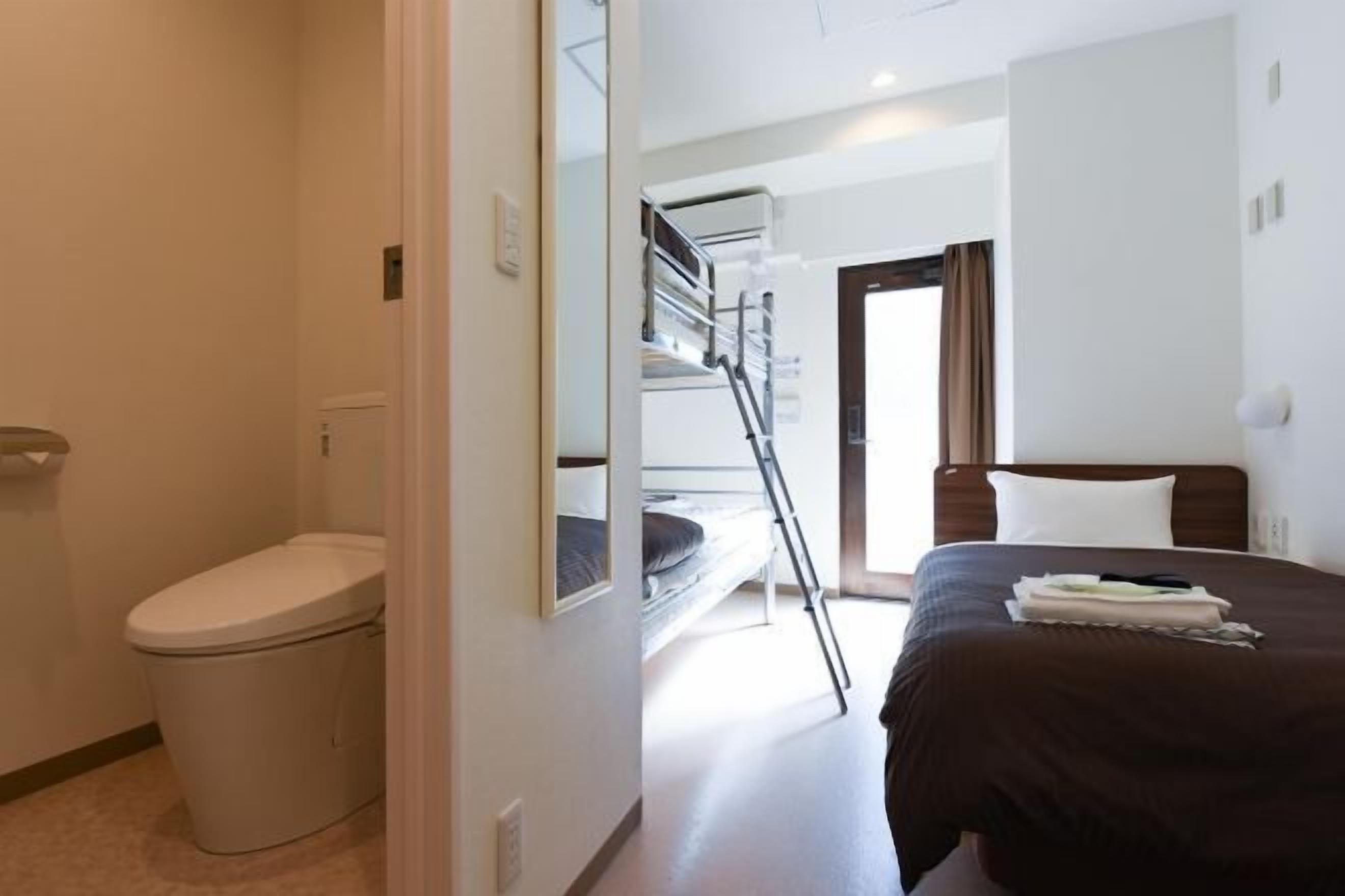 A hotel room with a bunk bed, a toilet, a bedside table with a lamp, and a window.