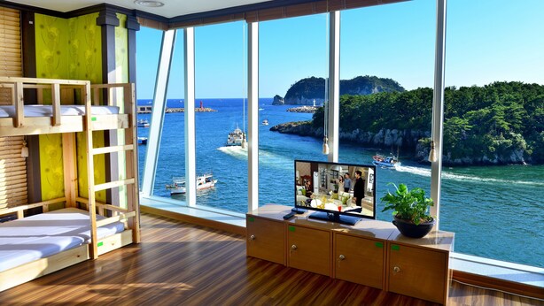 A room with a large window overlooking a coastal area, a bunk bed, a TV, and a cabinet with a plant. - Seom Guesthouse - Hostel (Seogwipo)