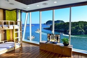 A room with a large window overlooking a coastal area, a bunk bed, a TV, and a cabinet with a plant. - Seom Guesthouse - Hostel (Seogwipo)