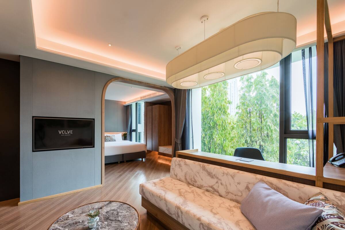 A modern hotel room with a marble sofa, a round coffee table with a plant, a bed with a TV mounted on the wall, and large windows offering a view of greenery.