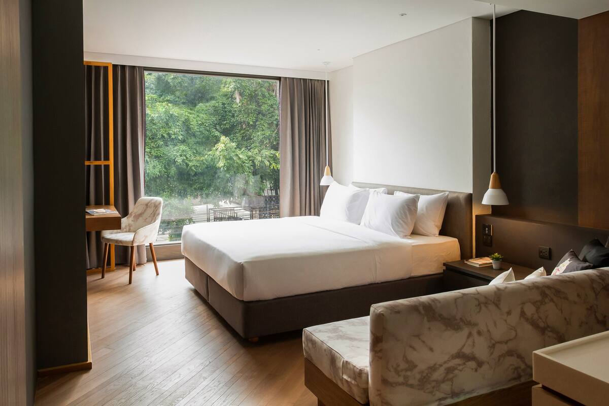 A modern hotel room with a large bed, a desk, and a chair. There is a view of greenery outside the window.