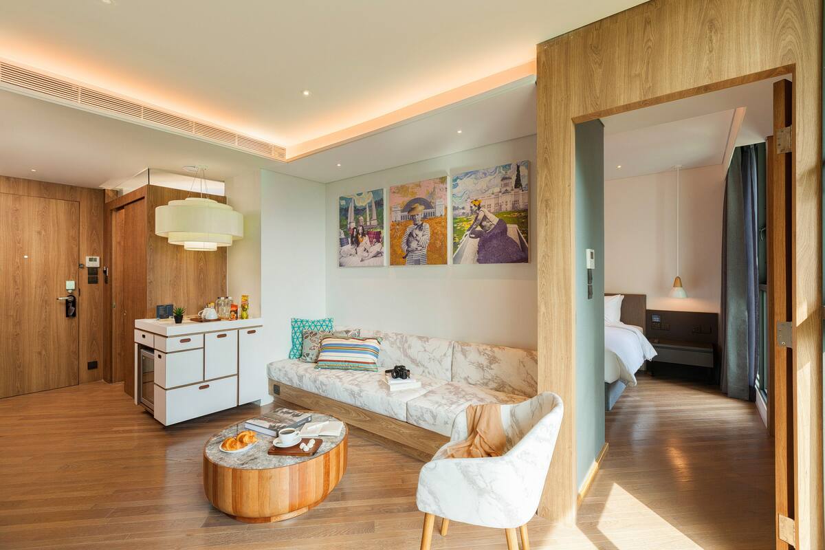 A modern hotel room with a wooden floor, a marble sofa, a round wooden coffee table, a white armchair, and a bed with a wooden headboard.