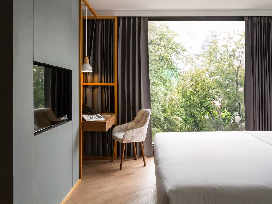 A hotel room with a bed, a desk with a chair, a telephone, and a view of trees and buildings through large windows.