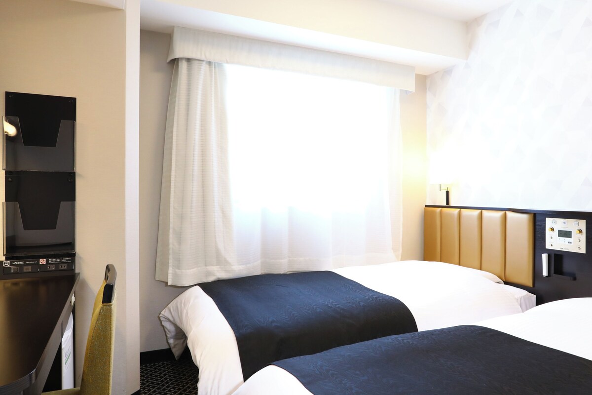 A hotel room with two beds, a TV, a window with a sheer curtain, and a control panel on the wall.
