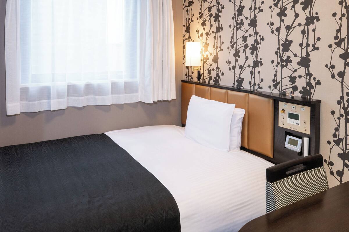 Quarto de hotel com cama, mesa de cabeceira com abajur, painel de controle e janela com cortinas finas.
