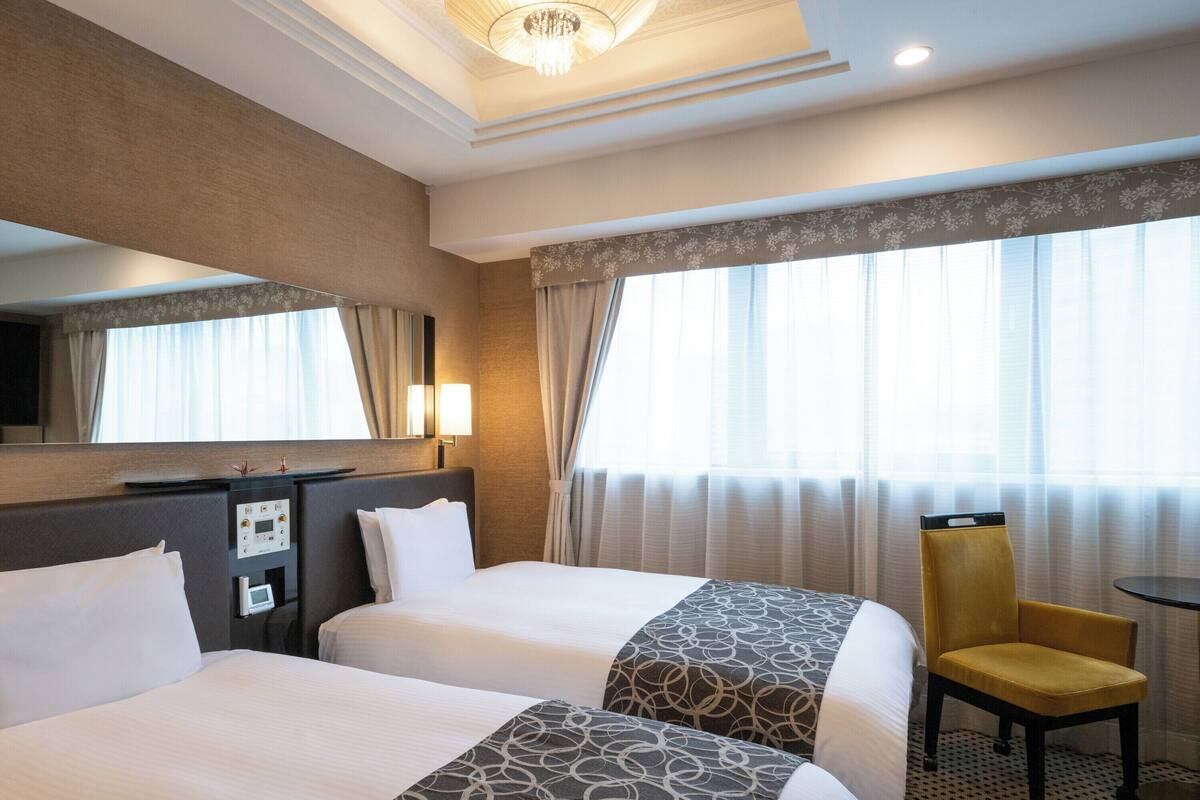 Quarto de hotel com duas camas, uma escrivaninha, uma cadeira, uma televisão e uma janela grande com cortinas.