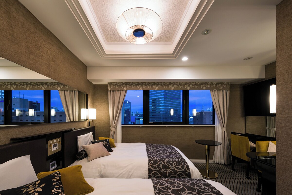 A hotel room with two beds, a desk, a chair, a TV, and a window with a city view.