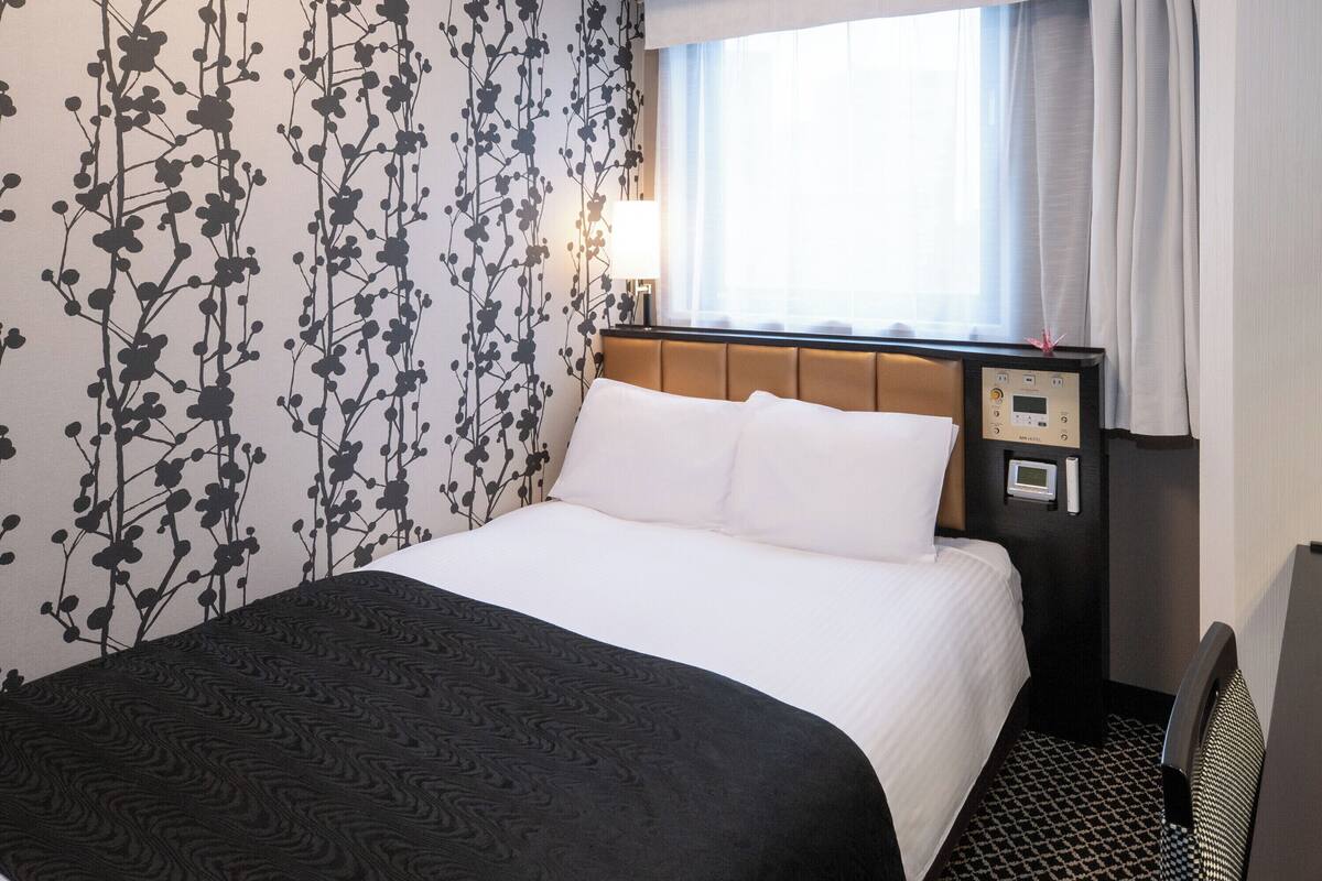 Quarto de hotel com cama, mesa de cabeceira, abajur, janela com cortinas e parede com padrão floral.