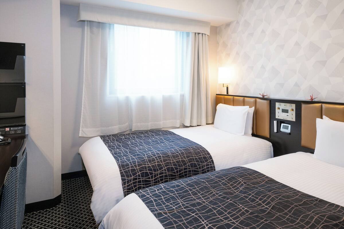 Quarto de hotel com duas camas, televisão, painel de controle e janela com cortinas.