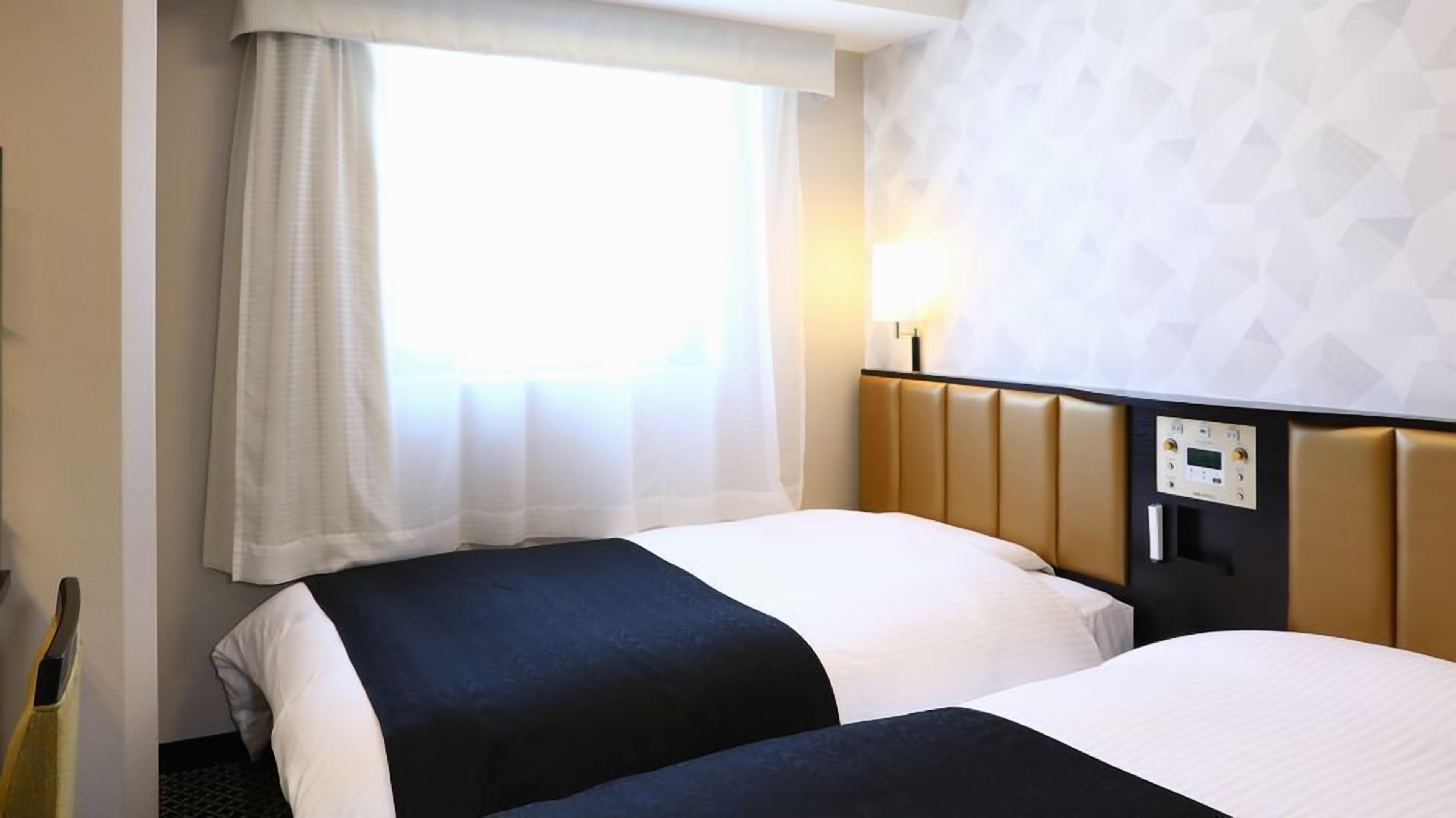 A hotel room with two beds, a window with sheer curtains, a wall-mounted control panel, and a patterned wall.