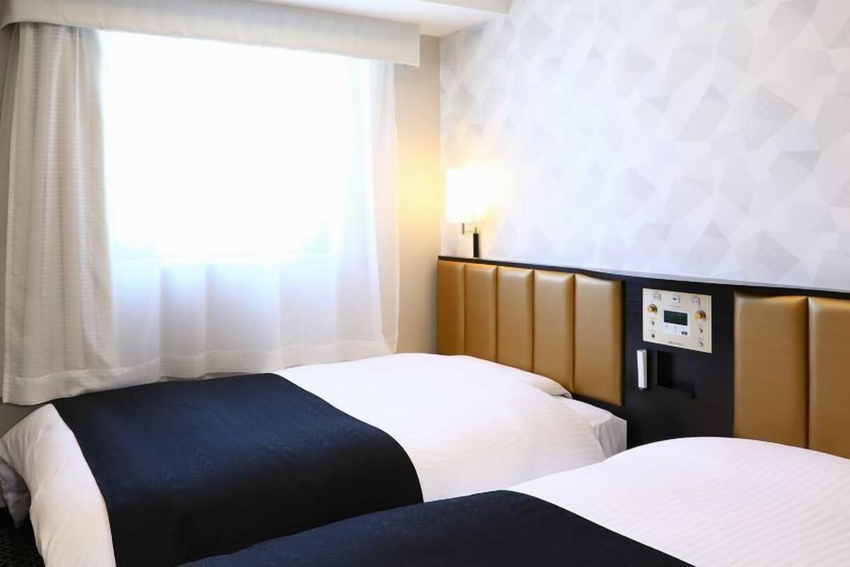 Quarto de hotel com duas camas, uma janela com cortinas finas, um painel de controle fixado na parede e um papel de parede estampado.
