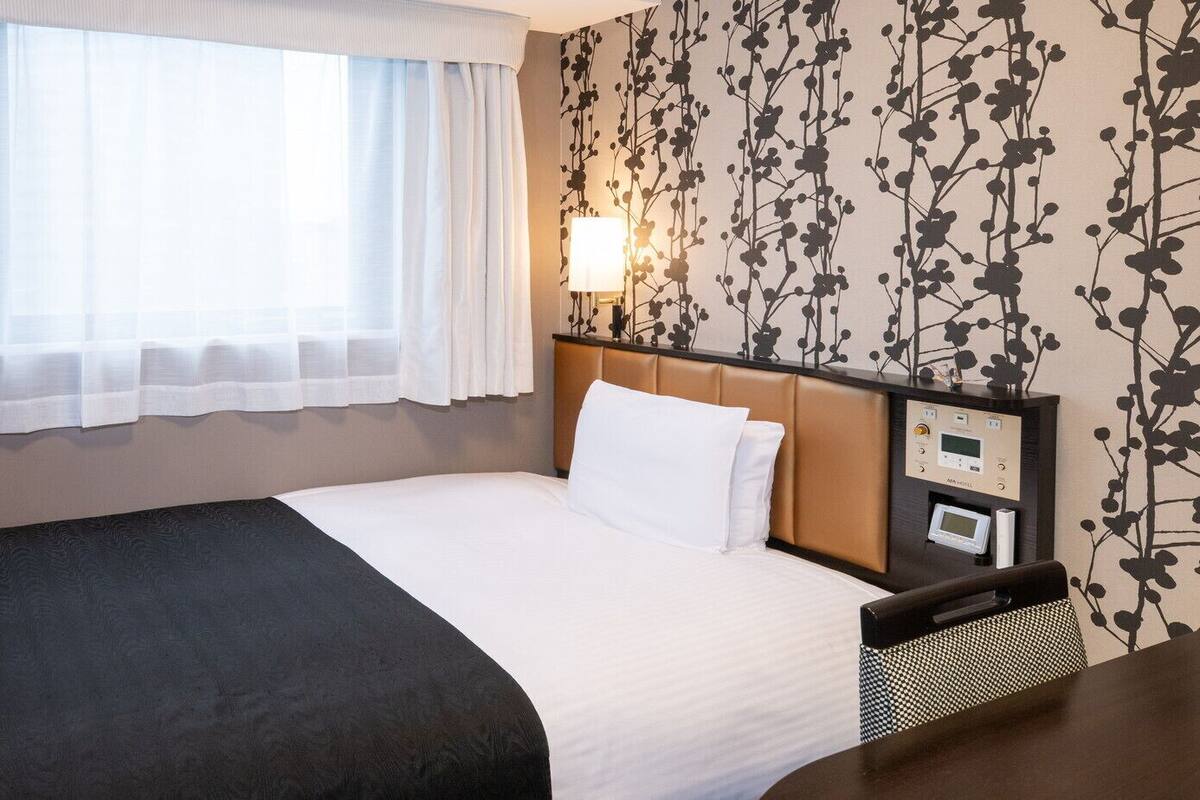 Quarto de hotel com cama, mesa de cabeceira, luminária e painel de controle.