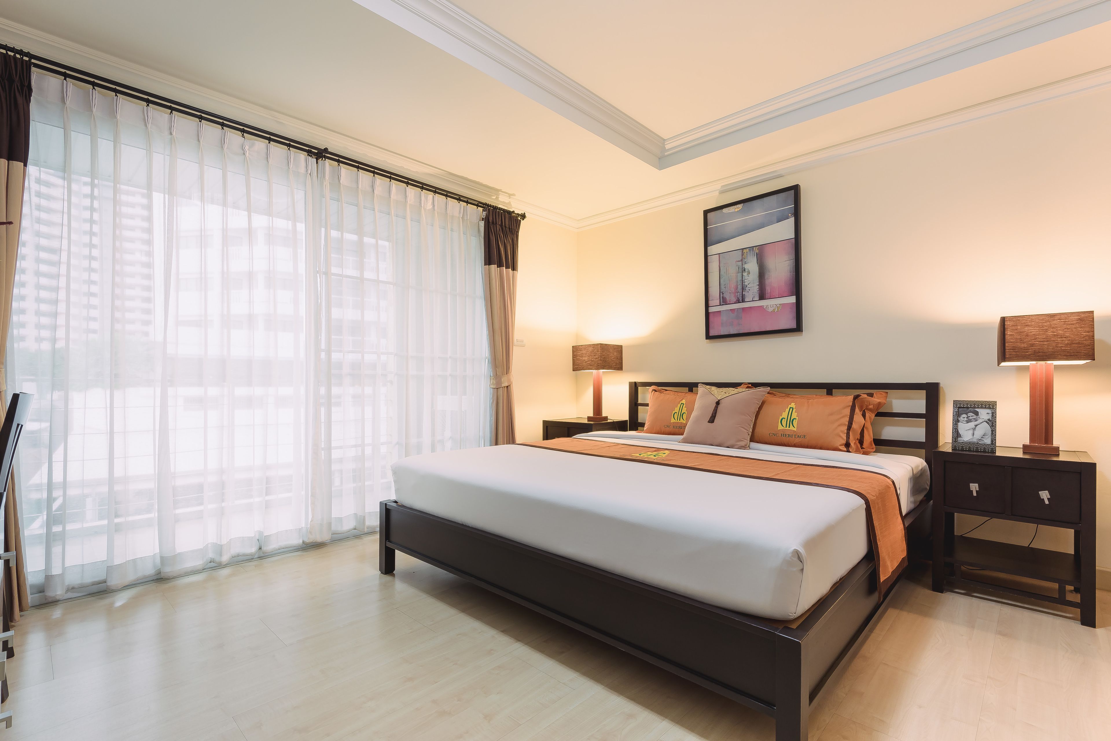 A hotel room with a large bed, two bedside tables with lamps, a painting on the wall, and sheer curtains on the window.